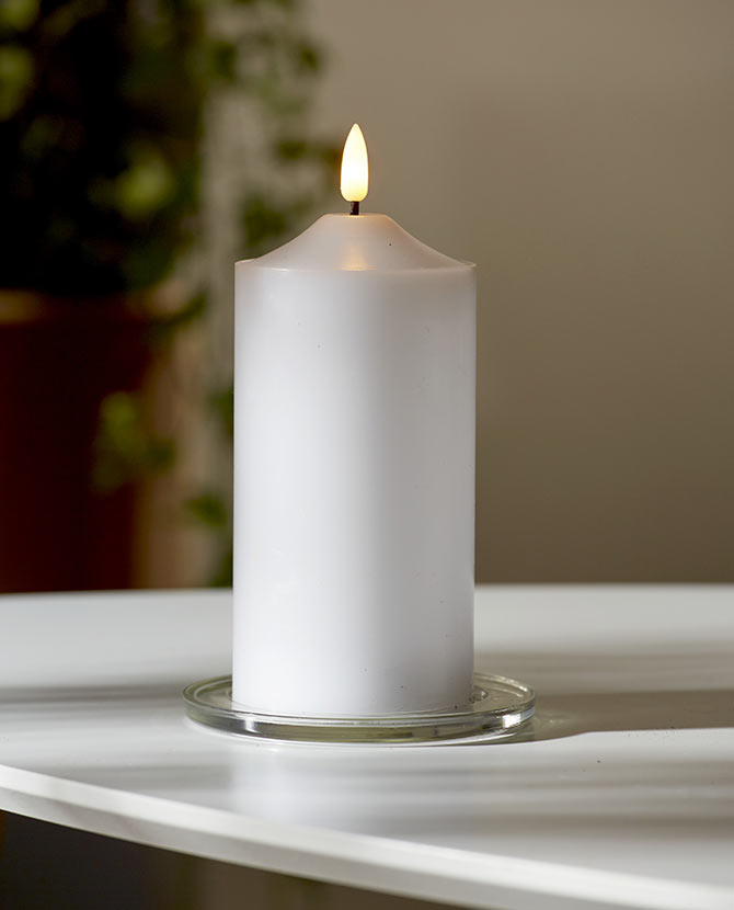 Valódi viaszból készült, fehér színű led gyertya meleg fehér fenyő led lánggal üveg tányéron, fehér színű asztalon, szürke fal előtt 