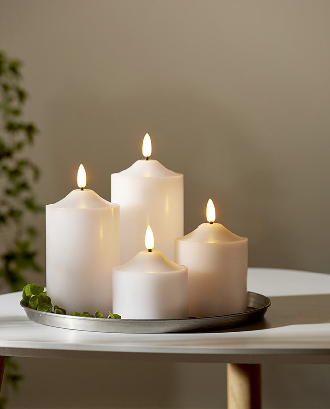 4 db különböző méretű, fehér színű viaszból készült led gyertya, meleg fehér fényű led lángokkal fém tálcán fehér modern asztalon, szürke fal előtt  