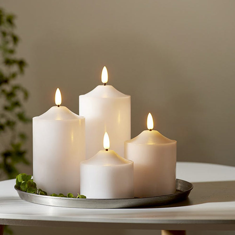 4 db különböző méretű, fehér színű viaszból készült led gyertya, meleg fehér fényű led lángokkal fém tálcán fehér modern asztalon, szürke fal előtt  