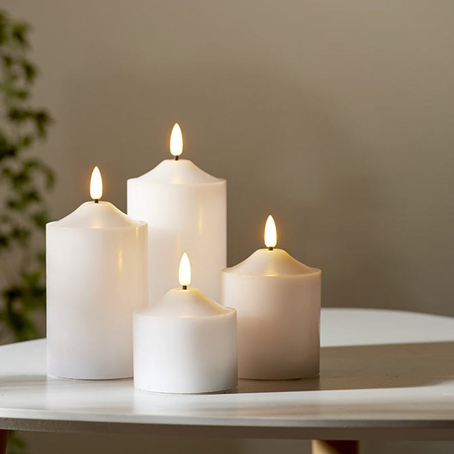 4 db különböző méretű, fehér színű viaszból készült led gyertya, meleg fehér fényű led lángokkal, fehér modern asztalon, szürke fal előtt