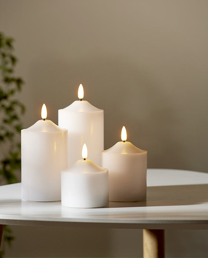 4 db különböző méretű, fehér színű viaszból készült led gyertya, meleg fehér fényű led lángokkal, fehér modern asztalon, szürke fal előtt