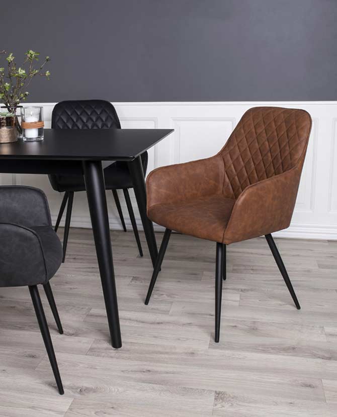 Modern formavilágú, skandináv stílusú, vintage barna színű, steppelt háttámlájú műbőr étkezőszék, fekete asztallal  laminált fa padlón, fehér kazettás lambéria előtt