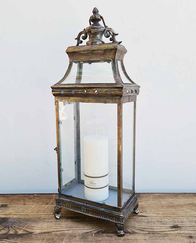 Barokkos megjelenésű, óarany színű, nagy fém lámpás, antikolt fa asztalon fehér gyertyával
