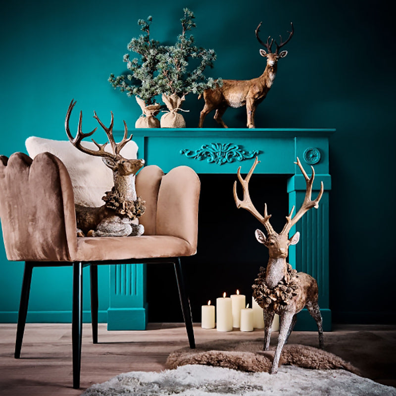 Erdei fenyőtoboz koszorúval díszített, nagyméretű karácsonyi szarvas figura , kék színű szobában, kék kandalló elött.