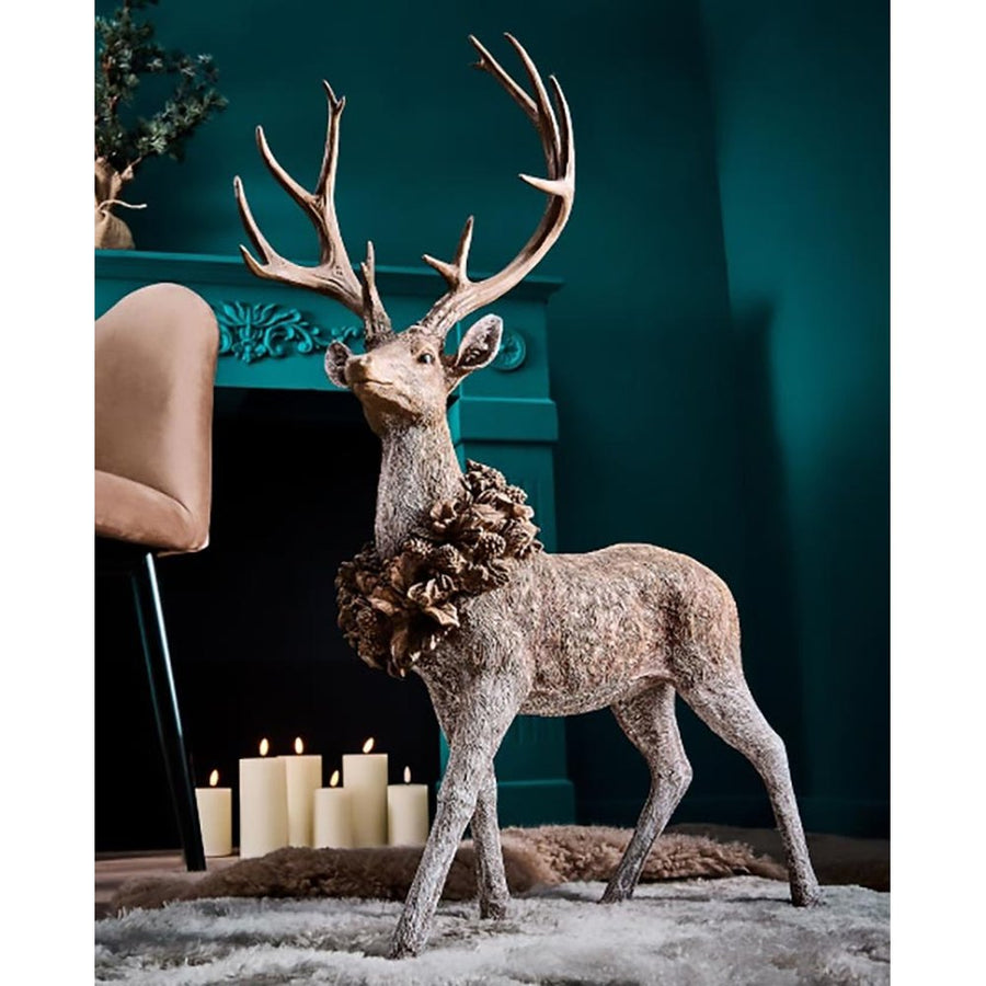 Erdei fenyőtoboz koszorúval díszített, nagyméretű karácsonyi szarvas figura, kék színű szobában, kék színű kandalló előtt.