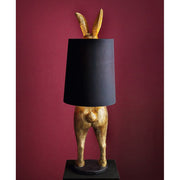 Aranyszínű, rejtőzködő nyuszi figurás, díjnyertes dizájn állólámpa, feketeszínű lámpaernyővel, bordó fal előtt.
