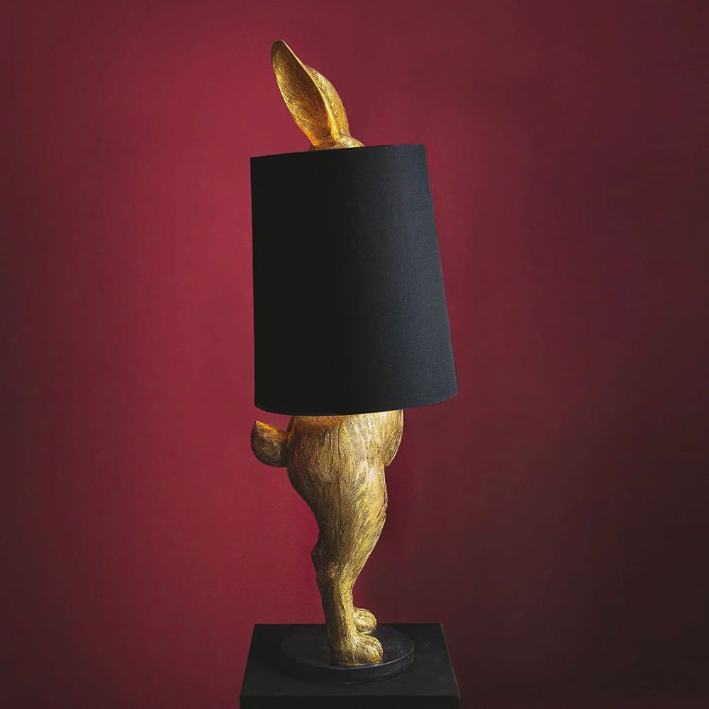 Aranyszínű, rejtőzködő nyuszi figurás, díjnyertes dizájn állólámpa, feketeszínű lámpaernyővel, bordó fal előtt.