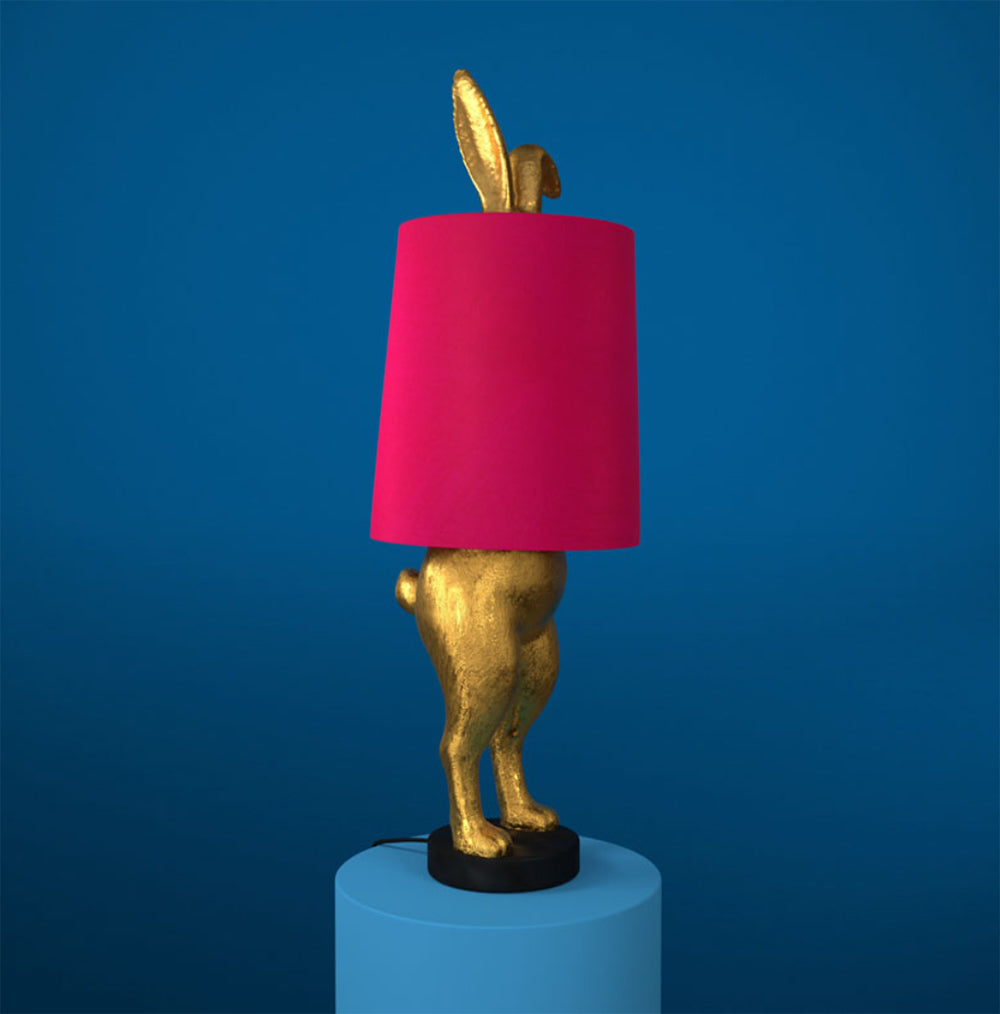 Aranyszínű, rejtőzködő nyuszi figurás, 74 cm magas, kortárs, glamour stílusú, díjnyertes dizájn asztali lámpa, pink színű lámpaernyővel, kékszínű háttér előtt.