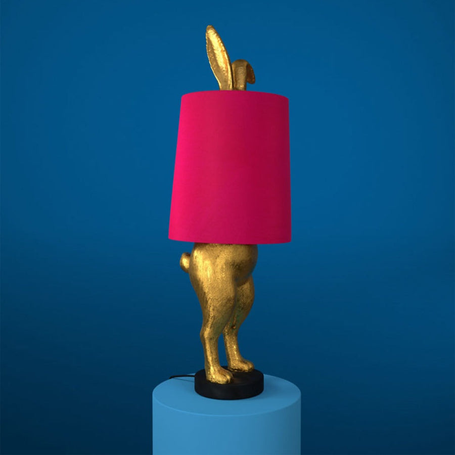 Aranyszínű, rejtőzködő nyuszi figurás, 74 cm magas, kortárs, glamour stílusú, díjnyertes dizájn asztali lámpa, pink színű lámpaernyővel, kékszínű háttér előtt.
