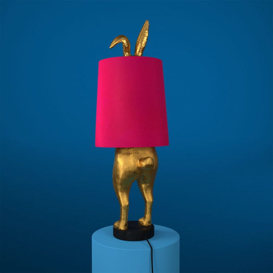Aranyszínű, rejtőzködő nyuszi figurás, 74 cm magas, kortárs, glamour stílusú, díjnyertes dizájn asztali lámpa, pink színű lámpaernyővel, kékszínű háttér előtt.
