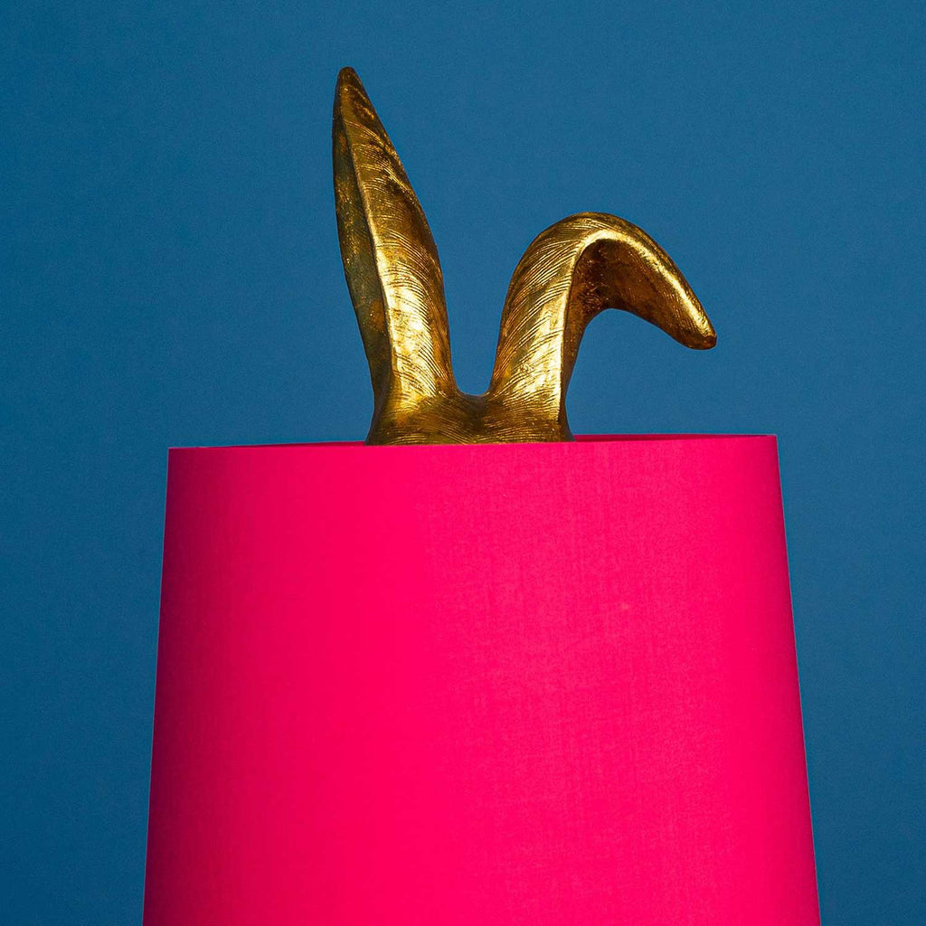 Aranyszínű, rejtőzködő nyuszi figurás, 74 cm magas, kortárs, glamour stílusú, díjnyertes dizájn asztali lámpa, pink színű lámpaernyővel, kékszínű háttér előtt.