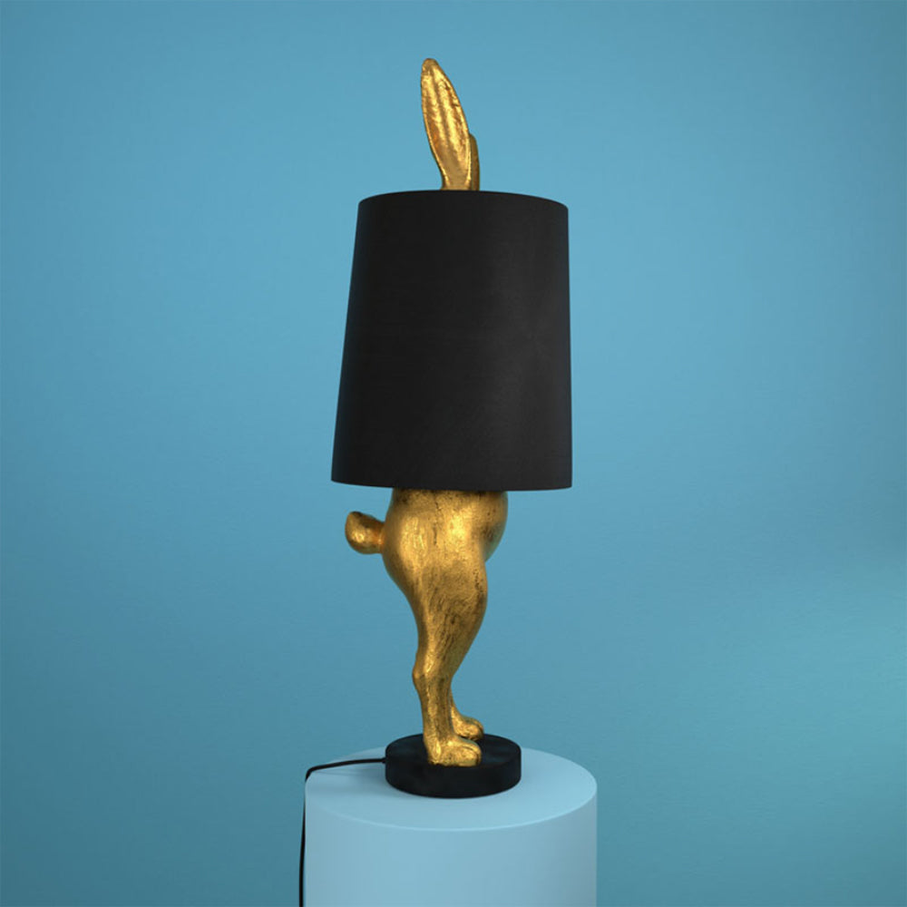 Aranyszínű, rejtőzködő nyuszi figurás, 74 cm magas, kortárs, glamour stílusú, díjnyertes dizájn asztali lámpa, feketeszínű lámpaernyővel, kék háttér előtt.