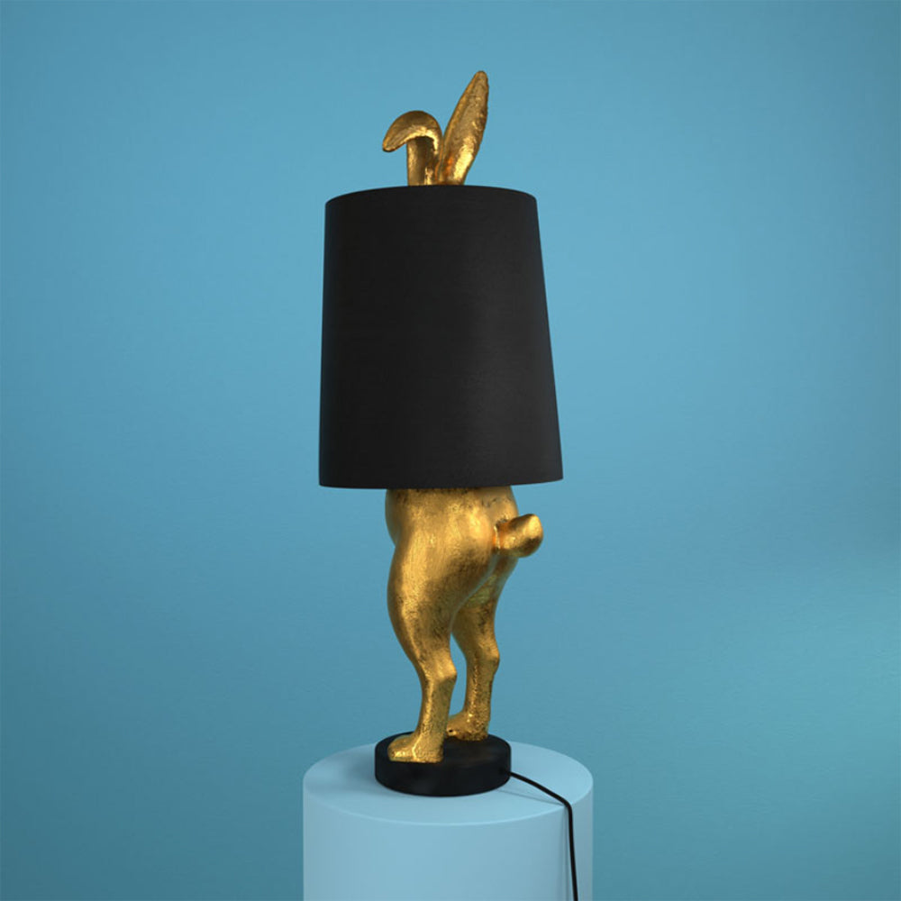Aranyszínű, rejtőzködő nyuszi figurás, 74 cm magas, kortárs, glamour stílusú, díjnyertes dizájn asztali lámpa, feketeszínű lámpaernyővel, kék háttér előtt.