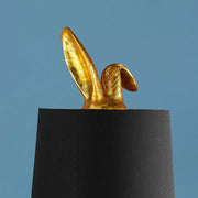 Aranyszínű, rejtőzködő nyuszi figurás, 74 cm magas, kortárs, glamour stílusú, díjnyertes dizájn asztali lámpa, feketeszínű lámpaernyővel, kék háttér előtt.
