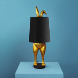 Aranyszínű, rejtőzködő nyuszi figurás, 74 cm magas, kortárs, glamour stílusú, díjnyertes dizájn asztali lámpa, feketeszínű lámpaernyővel, kék háttér előtt.