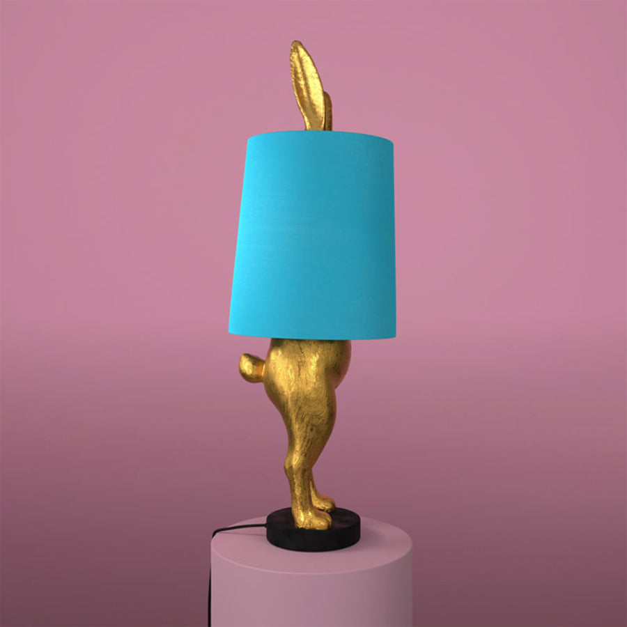 Aranyszínű, rejtőzködő nyuszi figurás, 74 cm magas, kortárs, glamour stílusú, díjnyertes dizájn asztali lámpa, türkizszínű lámpaernyővel, mályva színű fal előtt.