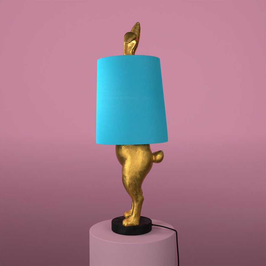 Aranyszínű, rejtőzködő nyuszi figurás, 74 cm magas, kortárs, glamour stílusú, díjnyertes dizájn asztali lámpa, türkizszínű lámpaernyővel, mályva színű fal előtt.