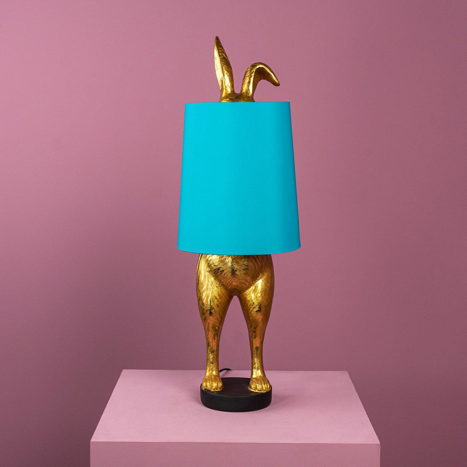 Aranyszínű, rejtőzködő nyuszi figurás, 74 cm magas, kortárs, glamour stílusú, díjnyertes dizájn asztali lámpa, türkizszínű lámpaernyővel, mályva színű fal előtt.