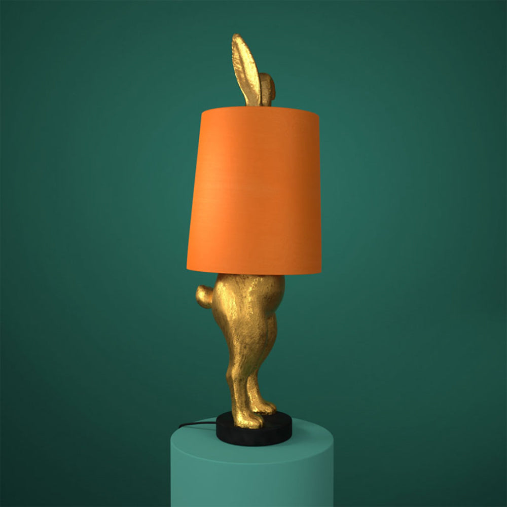 Aranyszínű, rejtőzködő nyuszi figurás, 74 cm magas, kortárs, glamour stílusú, díjnyertes dizájn asztali lámpa, narancsszínű lámpaernyővel, türkizzöld háttér előtt.