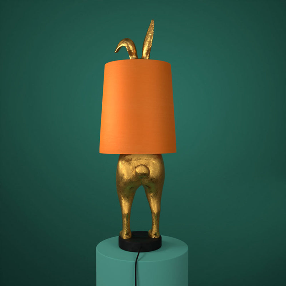 Aranyszínű, rejtőzködő nyuszi figurás, 74 cm magas, kortárs, glamour stílusú, díjnyertes dizájn asztali lámpa, narancsszínű lámpaernyővel, türkizzöld háttér előtt.