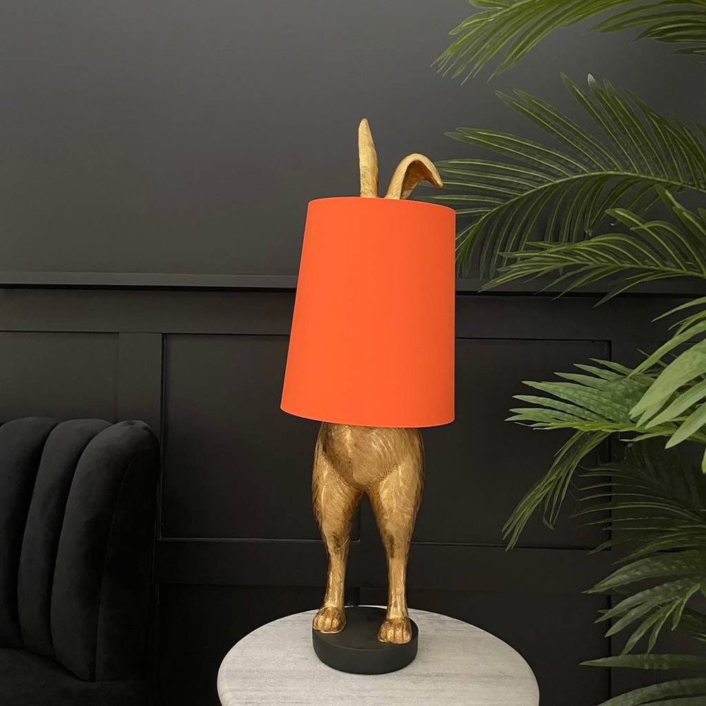 Aranyszínű, rejtőzködő nyuszi figurás, 74 cm magas, kortárs, glamour stílusú, díjnyertes dizájn asztali lámpa, narancsszínű lámpaernyővel, szürke fal előtt pálmával.