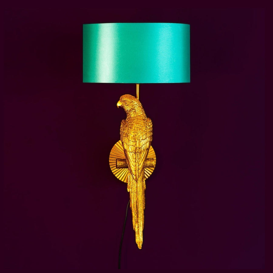 Aranyszínű papagáj figurával díszített, trópusi, glamour stílusú falilámpa, türkizszínű lámpaernyővel., lila háttér előtt.