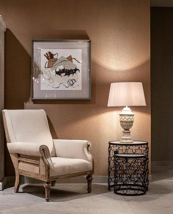 Prémium kategóriás, gyarmati stílusú, újrahasznosított trópusi fából készült, ekrü színű szövettel kárpitozott fotel barna fal előtt szürke padlón, koloniál asztali lámpával dohányzóasztalon