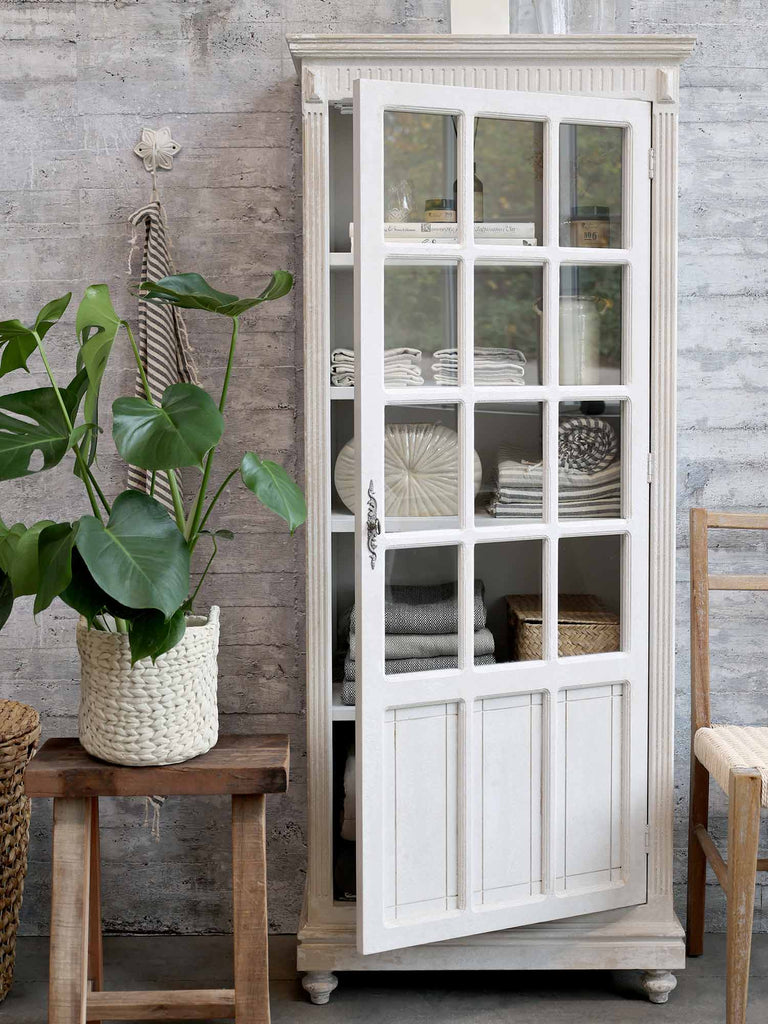 Provanszi stílusú, rusztikus felületű antik fehér színű vitrines tárolószekrény nyitott ajtóval, zöld növénnyel.