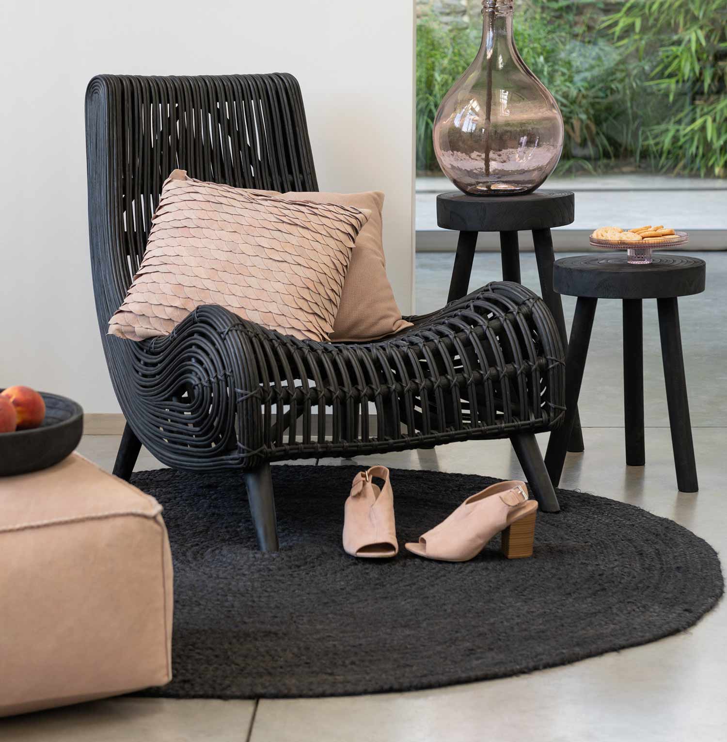Fekete színű rattan pihenő fotel teraszon, bézs párnákkal, női cipővel és vázával. 