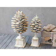 Kétféle méretű, antikolt krémszínű, fém toboz dísz szürke fal előtt, karácsonyi ajándékokkal.