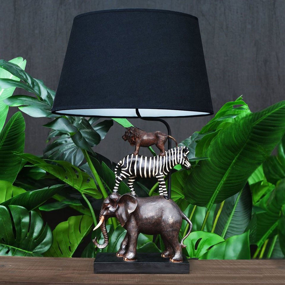 Szavannai állat figurás, 60 cm magas, trópusi stílusú asztali lámpa, zöld trópusi növények előtt.