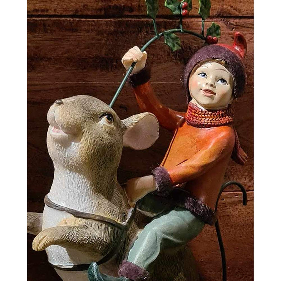 Barna színű erdei egér hátán utazó karácsonyi manó bordó ruhában és sállal, kezében magyallal barna színű deszkafal előtt