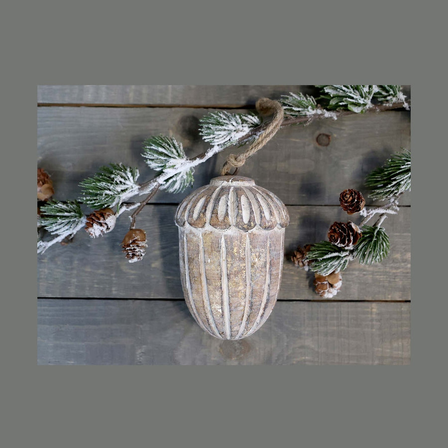 Vintage stílusú, mangófából faragott, 15 cm magas, antikolt felületű, tölgy makk formájú, kézműves karácsonyfadísz szürke deszkaasztalon havas fenyővel.
