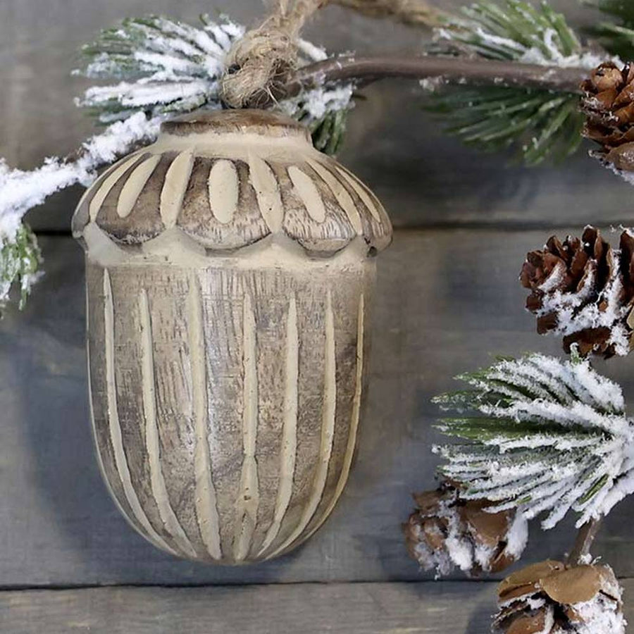 Vintage stílusú, mangófából faragott, 8 cm hosszú, antikolt felületű natúr színű, makk formájú, kézműves karácsonyfadísz szürke deszkaasztalon fenyőággal 