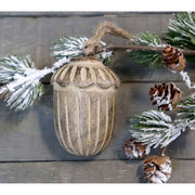 Vintage stílusú, mangófából faragott, 8 cm hosszú, antikolt felületű natúr színű, makk formájú, kézműves karácsonyfadísz szürke deszkaasztalon fenyőággal