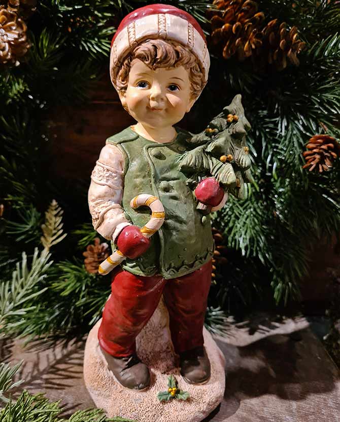 Vintage stílusú, 24,2 cm magas, tradicionális megjelenésű, karácsonyi téli sapkás kisfiú karácsonyfával a kezében zöld fenyőfa előtt