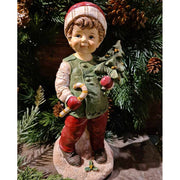 Vintage stílusú, 24,2 cm magas, tradicionális megjelenésű, karácsonyi téli sapkás kisfiú karácsonyfával a kezében zöld fenyőfa előtt