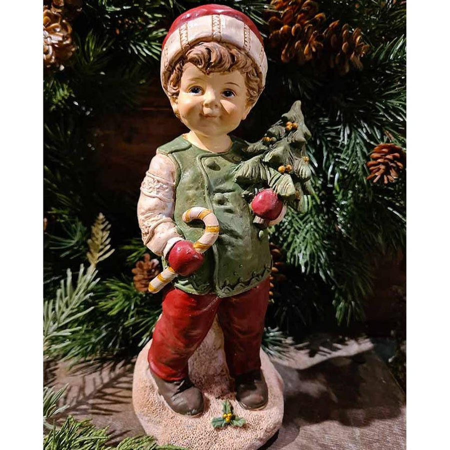 Vintage stílusú, 24,2 cm magas, tradicionális megjelenésű, karácsonyi téli sapkás kisfiú karácsonyfával a kezében zöld fenyőfa előtt