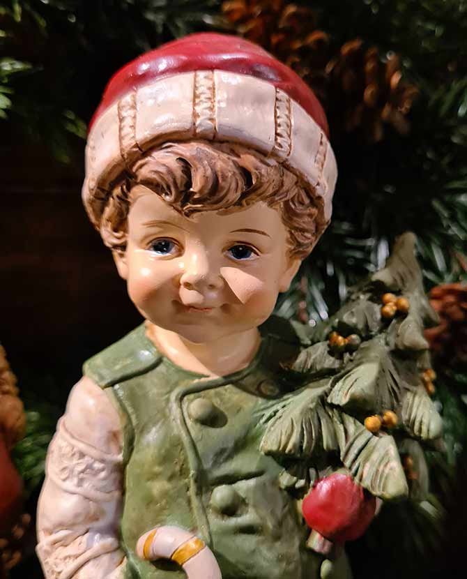 Vintage stílusú, 24,2 cm magas, tradicionális megjelenésű, karácsonyi téli sapkás kisfiú karácsonyfával a kezében zöld fenyőfa előtt