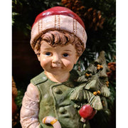 Vintage stílusú, 24,2 cm magas, tradicionális megjelenésű, karácsonyi téli sapkás kisfiú karácsonyfával a kezében zöld fenyőfa előtt