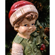 Vintage stílusú, 24,2 cm magas, tradicionális megjelenésű, karácsonyi téli sapkás kisfiú karácsonyfával a kezében zöld fenyőfa előtt