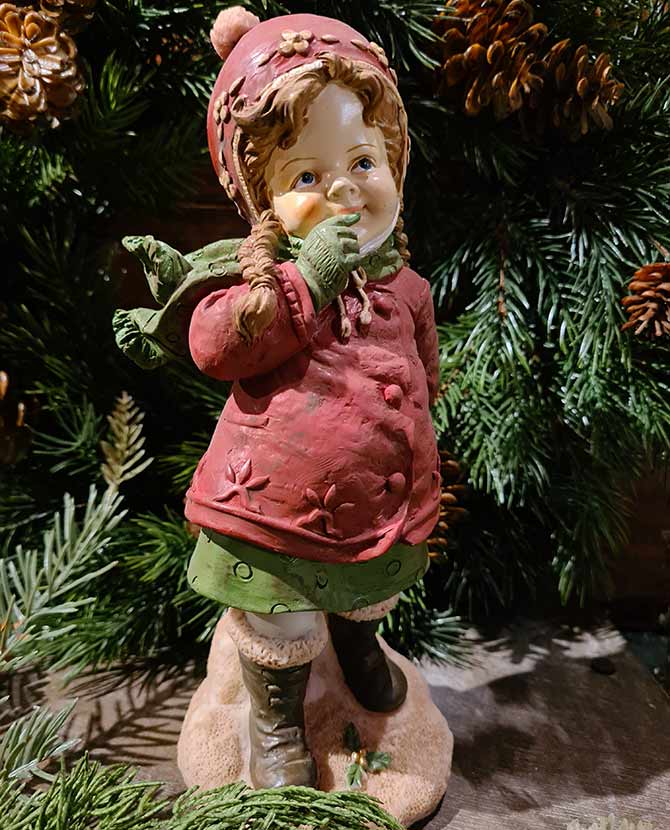 Vintage stílusú, 24,7 cm magas, tradicionális megjelenésű, karácsonyi téli sapkás kislány sállal a nyakában zöld fenyő előtt 