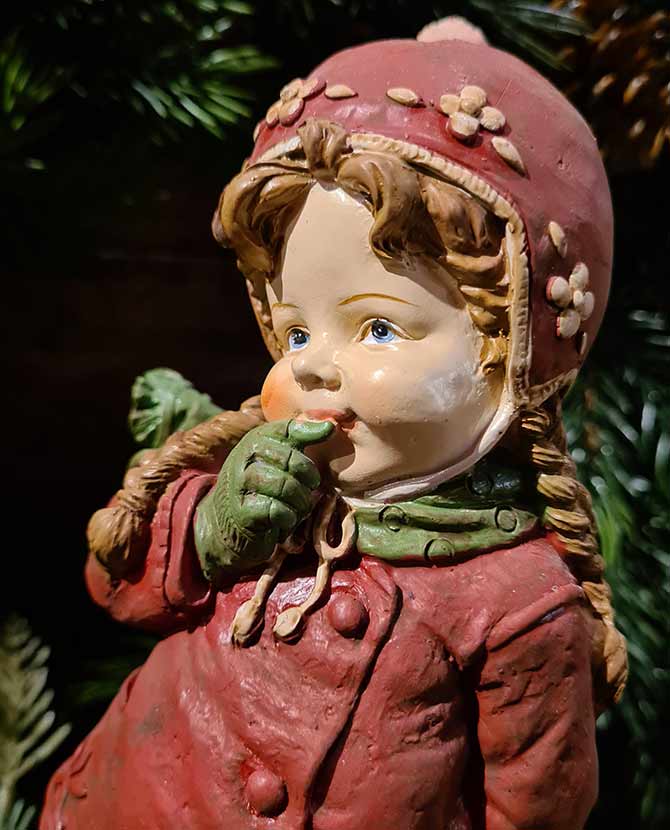 Vintage stílusú, 24,7 cm magas, tradicionális megjelenésű, karácsonyi téli sapkás kislány sállal a nyakában zöld fenyő előtt