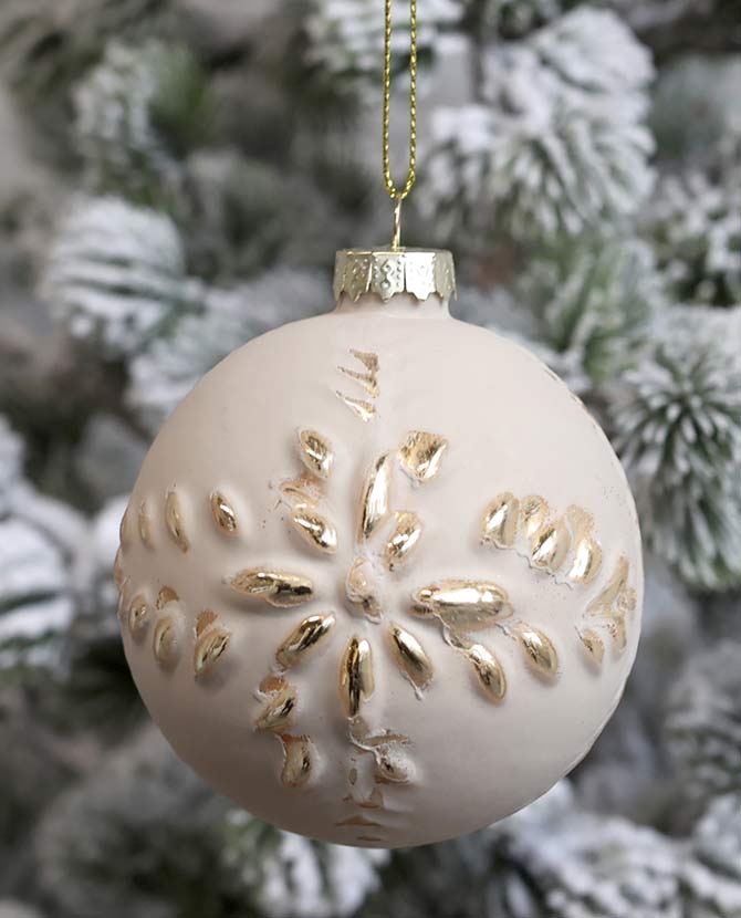 Vintage stílusú, aranyszínű növényi dombormintával díszített, gömb formájú, púderszínű üveg karácsonyfadísz, havas fenyőfán.