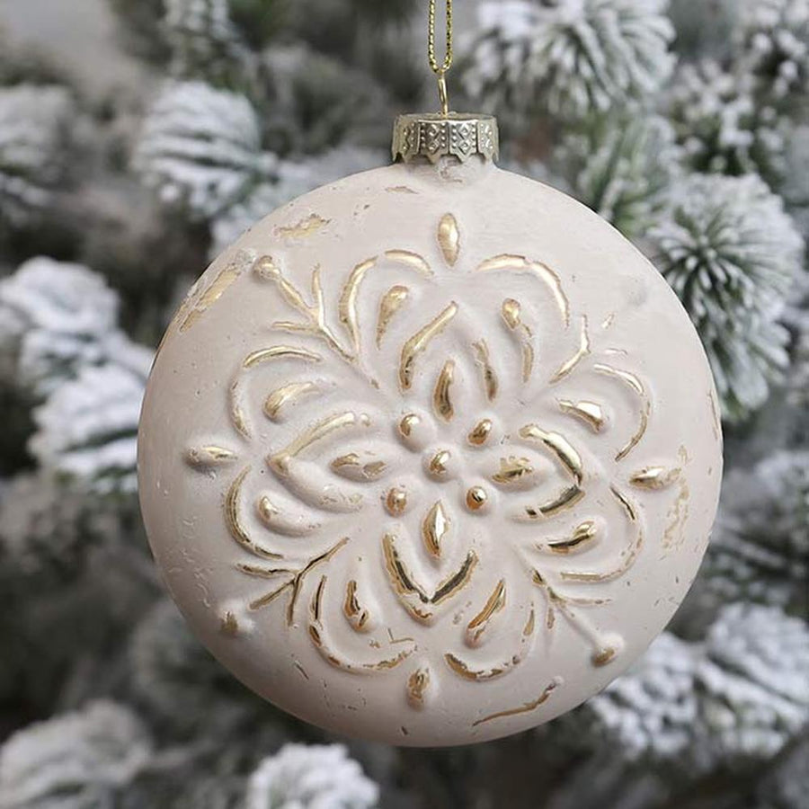 Vintage stílusú, aranyszínű növényi dombormintával díszített, gömb formájú, púderszínű üveg karácsonyfadísz, havas fenyőfán.